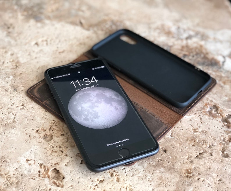 K&ouml;nnte beinhalten: Eine braune Leder-Handyh&uuml;lle mit einem schwarzen Telefon darin. Der Telefonbildschirm zeigt die Uhrzeit 11:34 und das Datum Mittwoch, 17. Juli. Die Handyh&uuml;lle hat eine Klappe, die &uuml;ber das Telefon geklappt wird.