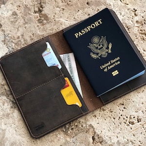Puede incluir: Un portapasaporte de cuero marr&oacute;n con un pasaporte azul oscuro y algunas tarjetas y efectivo en su interior. El pasaporte est&aacute; abierto, mostrando las palabras "Passport" y "United States of America". El portapasaporte est&aacute; abierto, revelando bolsillos para tarjetas y otros documentos.