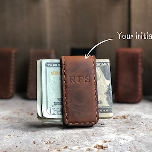 May include: A brown leather money clip with the initials "RFS" embossed on the front. The clip is holding a stack of US dollar bills.