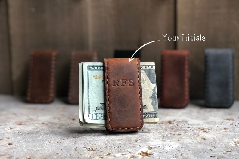 May include: A brown leather money clip with the initials "RFS" embossed on the front. The clip is holding a stack of US dollar bills.