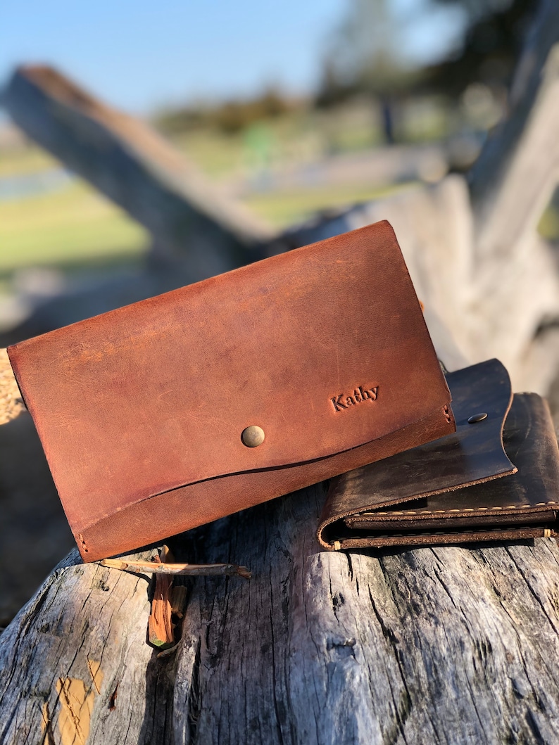 May include: Two brown leather wallets, one with the name "Kathy" embossed on the front. The wallets are sitting on a weathered wooden surface.