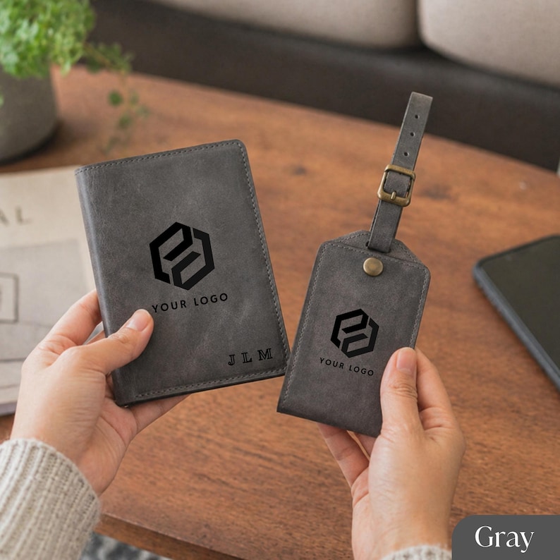 Puede incluir: Juego gris de funda para pasaporte y etiqueta para equipaje. Ambos art&iacute;culos presentan un logotipo negro y el texto "YOUR LOGO". El portapasaporte tambi&eacute;n tiene las iniciales "JLM" grabadas. La etiqueta de equipaje tiene una hebilla y una correa.