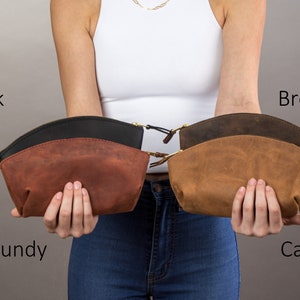 May include: Four leather cosmetic bags in different colors: burgundy, black, brown, and camel. The bags are held by a person's hands.