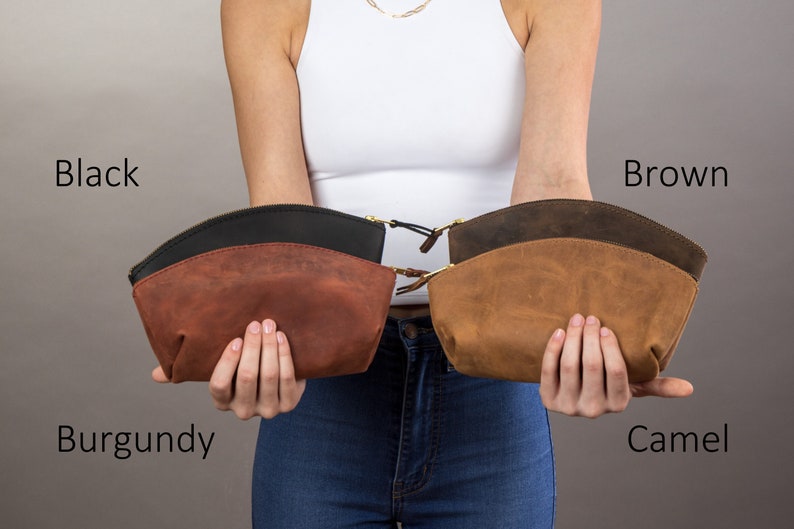 May include: Four leather cosmetic bags in different colors: burgundy, black, brown, and camel. The bags are held by a person's hands.