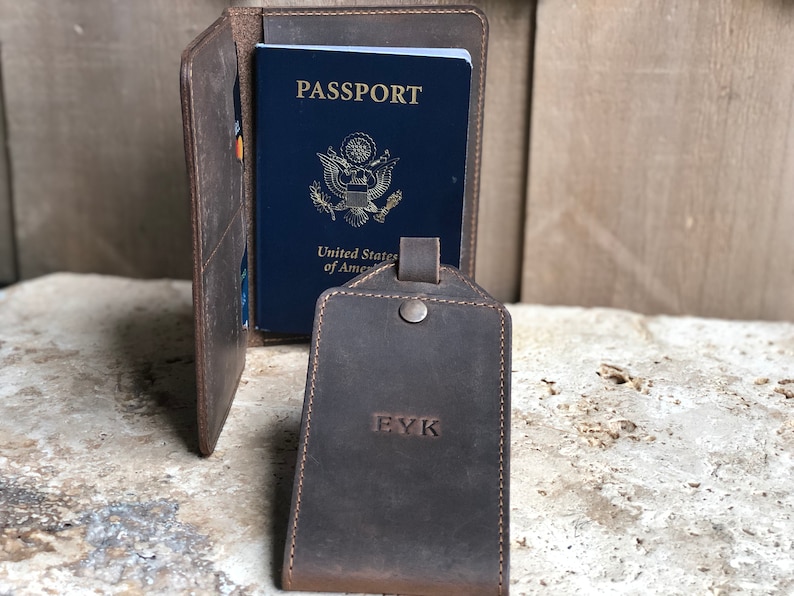 Puede incluir: Un portapasaporte de cuero marr&oacute;n con un pasaporte azul en su interior. Una etiqueta de equipaje a juego con las iniciales "EYK" est&aacute; al frente. El portapasaporte tiene ranuras para tarjetas. Los art&iacute;culos est&aacute;n sobre una superficie texturizada.