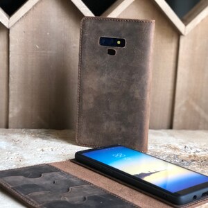 K&ouml;nnte beinhalten: Eine braune Leder-Handyh&uuml;lle mit integriertem Portemonnaie. Das Etui hat mehrere Kartenf&auml;cher und ein Fach f&uuml;r Bargeld. Die Handyh&uuml;lle ist ge&ouml;ffnet und ein schwarzes Smartphone befindet sich darin.