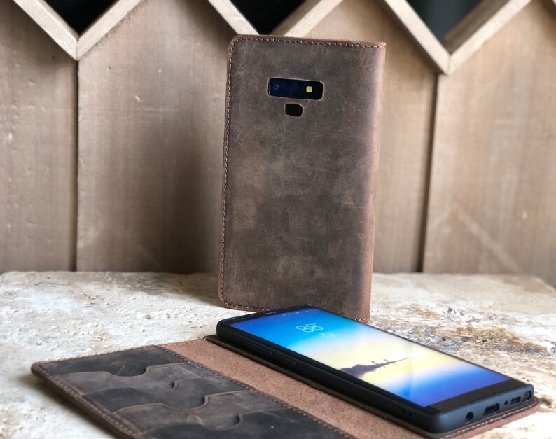 K&ouml;nnte beinhalten: Eine braune Leder-Handyh&uuml;lle mit integriertem Portemonnaie. Das Etui hat mehrere Kartenf&auml;cher und ein Fach f&uuml;r Bargeld. Die Handyh&uuml;lle ist ge&ouml;ffnet und ein schwarzes Smartphone befindet sich darin.
