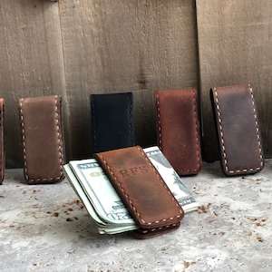 May include: Eight leather money clips in various colors, including brown, black, and gray. One clip has the initials "RFS" embossed on it and is holding a stack of bills.