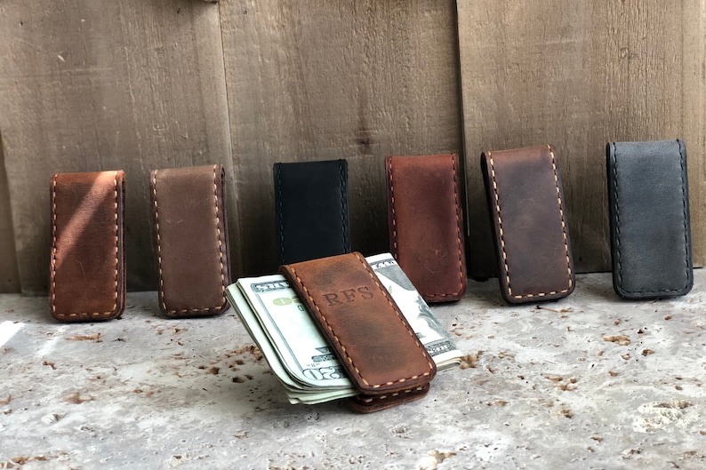 May include: Eight leather money clips in various colors, including brown, black, and gray. One clip has the initials "RFS" embossed on it and is holding a stack of bills.
