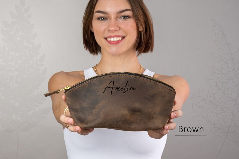 May include: A brown leather makeup bag with a zipper closure. The bag is personalized with the name "Amelia" in black script.