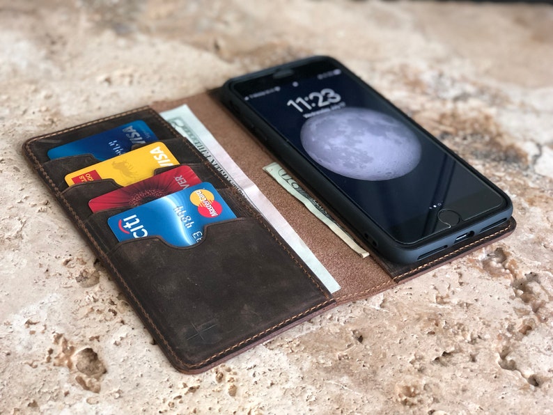 K&ouml;nnte beinhalten: Ein braunes Leder-Handy-Etui mit Steckpl&auml;tzen f&uuml;r Kreditkarten und Bargeld. Das Etui ist ge&ouml;ffnet und ein schwarzes Smartphone mit einem Mond-Hintergrundbild ist sichtbar. Die Uhrzeit auf dem Telefon ist 11:23.