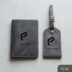 May include: Gray leather passport holder and luggage tag set. Both items feature a black logo and the text "YOUR LOGO". The luggage tag has a buckle and strap. The set is designed for travel.