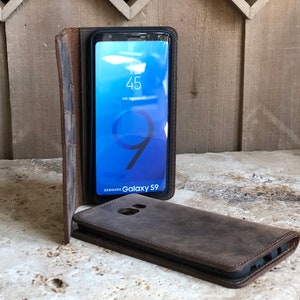 K&ouml;nnte beinhalten: Eine braune Leder-Handyh&uuml;lle mit einer Klappe, die sich &uuml;ber den Bildschirm eines Samsung Galaxy S9-Handys klappen l&auml;sst. Das Handy zeigt die Uhrzeit und das Datum auf dem Bildschirm an.