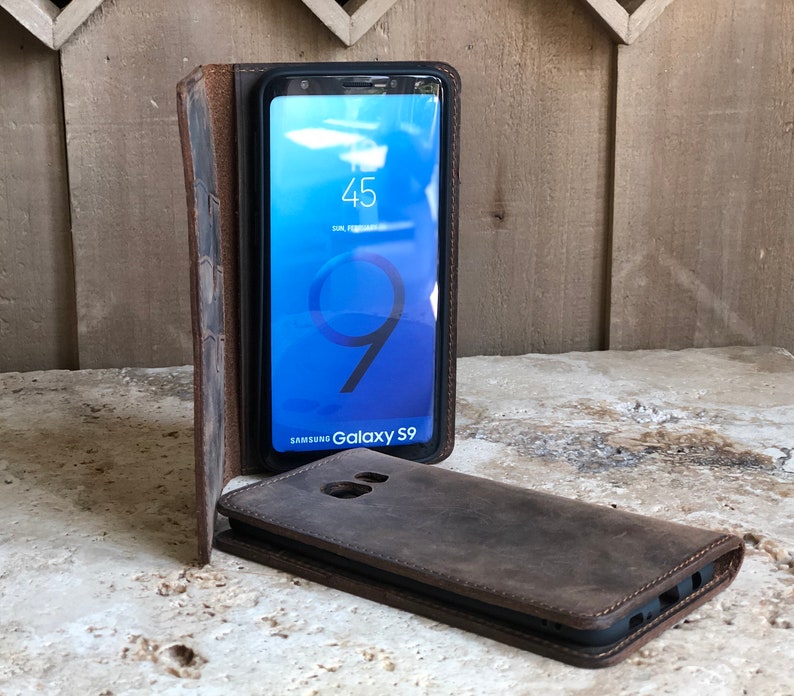 K&ouml;nnte beinhalten: Eine braune Leder-Handyh&uuml;lle mit einer Klappe, die sich &uuml;ber den Bildschirm eines Samsung Galaxy S9-Handys klappen l&auml;sst. Das Handy zeigt die Uhrzeit und das Datum auf dem Bildschirm an.