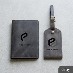 Puede incluir: Juego de viaje de cuero gris que incluye un tarjetero para pasaporte y una etiqueta para equipaje. Ambos art&iacute;culos presentan un logotipo negro y el texto "YOUR LOGO". La etiqueta para equipaje tiene una hebilla y una correa.