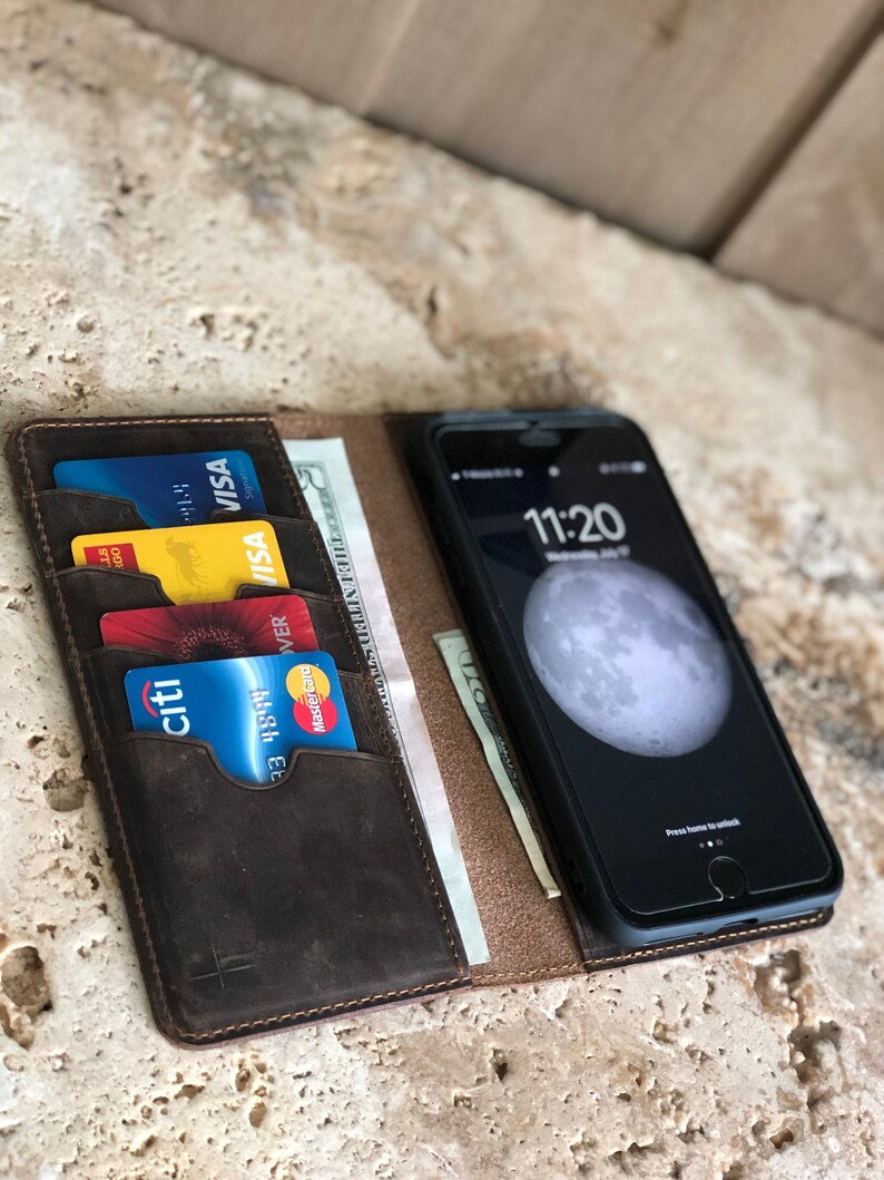 K&ouml;nnte beinhalten: Eine braune Ledergeldb&ouml;rse mit einem integrierten Handy-Etui. Die Geldb&ouml;rse hat mehrere Kartenf&auml;cher und ein Fach f&uuml;r Bargeld. Das Handy-Etui hat einen schwarzen Bildschirm mit einem wei&szlig;en Mond und die Uhrzeit 11:20 angezeigt.