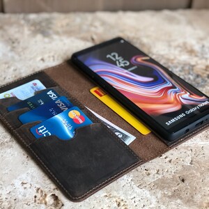 K&ouml;nnte beinhalten: Eine braune Leder-Wallet-H&uuml;lle mit einem Telefon und Kreditkarten im Inneren. Das Telefon ist ein Samsung Galaxy Note9 mit einem bunten abstrakten Bildschirmschoner. Die H&uuml;lle hat mehrere Kartenf&auml;cher und ein Fach f&uuml;r Bargeld.