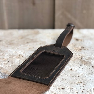 May include: A dark brown leather luggage tag with a rectangular window for an ID. The tag has a stitched border and a strap for attaching to luggage. The leather has a distressed appearance.