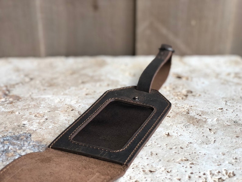 May include: A dark brown leather luggage tag with a rectangular window for an ID. The tag has a stitched border and a strap for attaching to luggage. The leather has a distressed appearance.