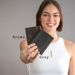 May include: Two leather wallets, one brown and one grey, held in a person's hand. The wallets are closed and have a simple design.