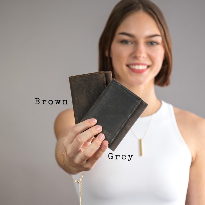 May include: Two leather wallets, one brown and one grey, held in a person's hand. The wallets are closed and have a simple design.