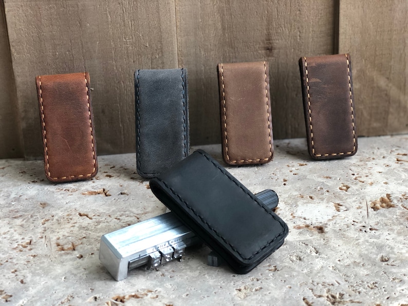 May include: Five leather money clips in different colors: brown, gray, brown, brown, and black. The clips are stitched with a contrasting thread.