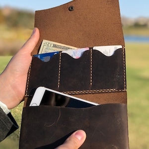 May include: A brown leather wallet with multiple card slots and a large pocket for a phone. The wallet is open and has a twenty dollar bill and a credit card inside.