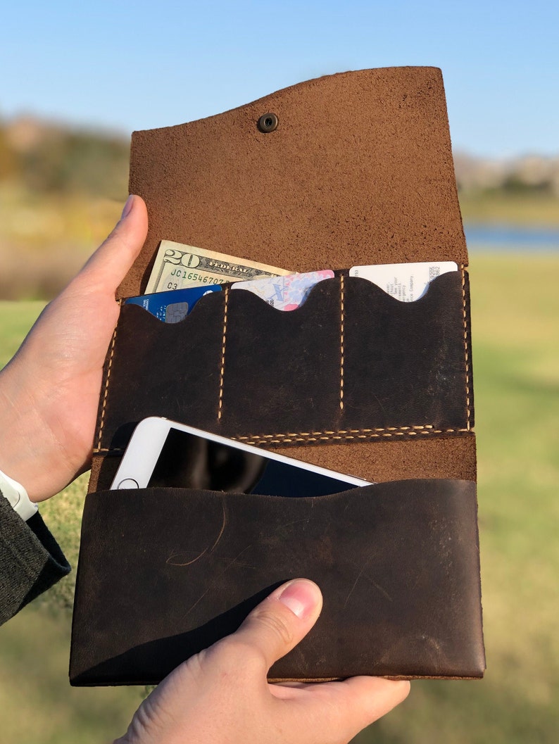 May include: A brown leather wallet with multiple card slots and a large pocket for a phone. The wallet is open and has a twenty dollar bill and a credit card inside.