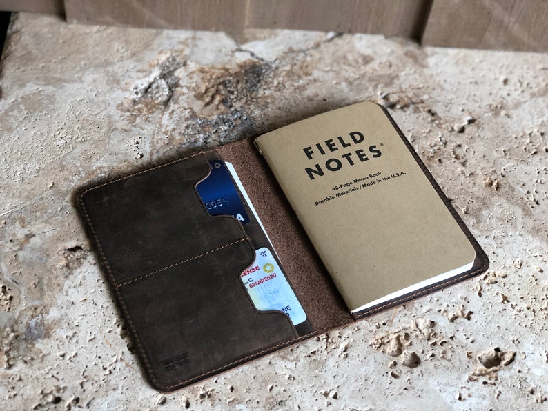 May include: Open brown leather wallet with a Field Notes memo book. The wallet has slots for cards and a driver's license. The Field Notes book is tan with black text. The wallet is on a textured surface.