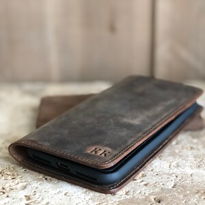 K&ouml;nnte beinhalten: Eine braune Leder-Handyh&uuml;lle mit Klappverschluss und den eingepr&auml;gten Initialen "KK" auf der Vorderseite.
