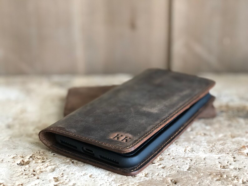 K&ouml;nnte beinhalten: Eine braune Leder-Handyh&uuml;lle mit Klappverschluss und den eingepr&auml;gten Initialen "KK" auf der Vorderseite.