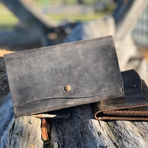 May include: Two leather wallets, one dark gray and one brown, with a brass button on the gray wallet. The wallets are sitting on a weathered wooden surface.