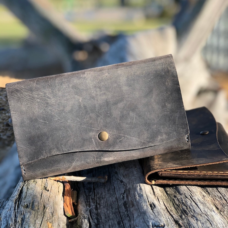 May include: Two leather wallets, one dark gray and one brown, with a brass button on the gray wallet. The wallets are sitting on a weathered wooden surface.