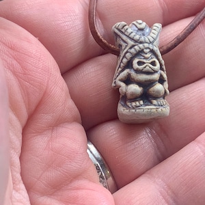 K&ouml;nnte beinhalten: Ein kleiner, cremefarbener Tiki-Anh&auml;nger mit einer braunen Lederschnur. Der Anh&auml;nger zeigt eine geschnitzte Figur mit detailliertem Kopfschmuck und einer hockenden Pose. Der Tiki hat ein verwittertes Aussehen, das auf eine gealterte oder Vintage-&Auml;sthetik hindeutet.