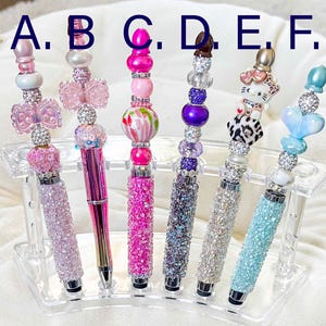 May include: A collection of six decorative pens with various embellishments. Each pen features a jeweled barrel and a beaded top with charms, including bows, butterflies, and a heart. The pens are displayed in a clear acrylic holder.