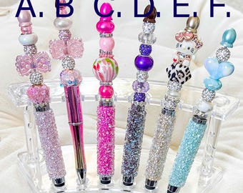 Op de afbeelding: Een verzameling van zes decoratieve pennen met verschillende versieringen. Elke pen heeft een met juwelen bezette schacht en een kralen top met bedels, waaronder strikken, vlinders en een hart. De pennen worden getoond in een heldere acrylhouder.
