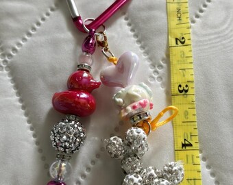 Könnte beinhalten: Ein herzförmiger, pinker Karabiner-Schlüsselanhänger mit zwei Perlensträngen. Ein Strang hat eine rote Ente, eine Strasskugel und eine pinke Herzperle. Der andere hat ein weißes Herz, einen mit Strass besetzten Pudelanhänger und eine gelbe Kordel. Ein gelbes Maßband zeigt eine Länge von ca. 12,7 cm.