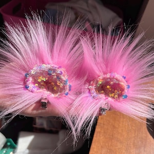 Peut inclure: Deux pinces à cheveux en fausse fourrure rose. Chaque pince présente un groupe central de paillettes de différentes couleurs, entouré d'un anneau de paillettes bleues et irisées. Les pinces sont fixées à des fermoirs en métal argenté.