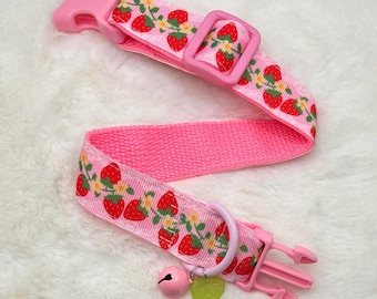 May include: A pink pet collar featuring a repeating pattern of red strawberries with green leaves and small yellow flowers. The collar includes a pink plastic side-release buckle, an adjustable slider, a metal D-ring, and a small pink bell charm.