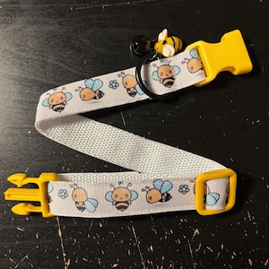 May include: White pet collar with a repeating pattern of cartoon bees in black, yellow, and blue. The collar has yellow plastic buckles and a small bee charm with a black bell.