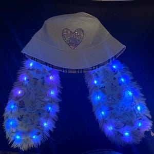 Puede incluir: Sombrero de pescador blanco con un corazón de lentejuelas y solapas largas y esponjosas con estampado. Las solapas están iluminadas con luces LED azules, creando un accesorio caprichoso y llamativo.