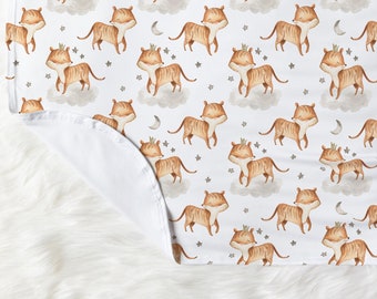 May include: A white baby blanket with a repeating pattern of brown tigers wearing crowns, white clouds, and silver stars and crescent moons.