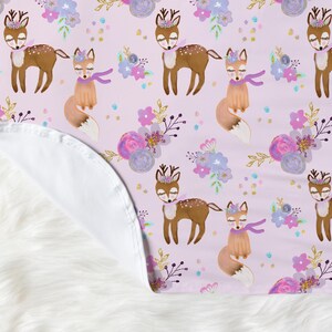 May include: A pink baby blanket with a repeating pattern of deer and foxes with floral accents. The deer and foxes are wearing crowns and the flowers are in shades of purple, pink, and blue.
