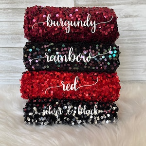 May include: A stack of four rolls of sequin fabric in different colors. The top roll is burgundy, the second roll is rainbow, the third roll is red, and the bottom roll is silver and black.