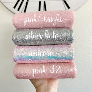 May include: Four rolls of fabric in shades of pink and silver are stacked. The top roll is light pink with the text "pink / bright". The second roll is silver with the text "silver holo". The third roll is pink with iridescent sequins and the text "unicorn". The bottom roll is pink with the text "pink 32".