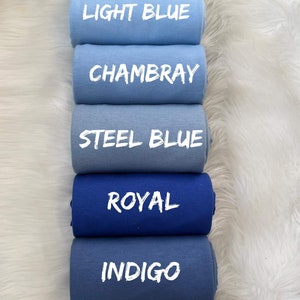May include: A stack of seven fabric rolls in shades of blue, from light to dark. The colors are labeled: New Light Blue, Light Blue, Chambray, Steel Blue, Royal, Indigo, and Navy. The rolls are arranged on a white, fluffy surface.