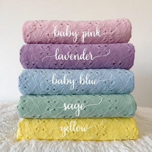May include: A stack of five eyelet fabric rolls in pastel colors: baby pink, lavender, baby blue, sage, and yellow. Each roll has a different color and is labeled with its respective color name in cursive script.
