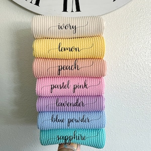 May include: Stack of seven waffle knit fabric swatches in various pastel colors, including ivory, lemon, peach, pastel pink, lavender, blue powder, and sapphire. Each swatch has the color name written in cursive on the fabric.