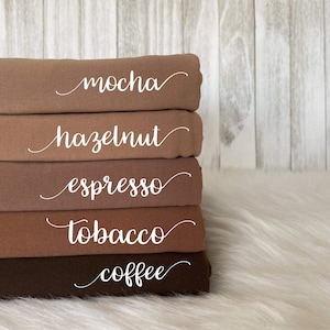 May include: Five fabric swatches stacked on top of each other. The swatches are different shades of brown, from light to dark. The swatches are labeled with the following names: mocha, hazelnut, espresso, tobacco, and coffee.