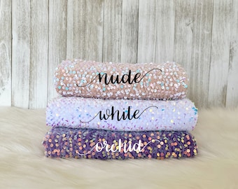 May include: Three rolls of iridescent sequin fabric in nude, white, and orchid colors. The fabric is stacked on a white faux fur surface.